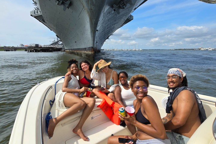Group of friends smiling on a boat near a large ship under a sunny sky.