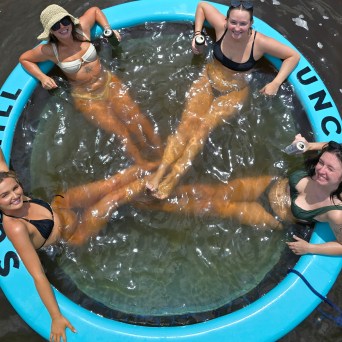 Four women in swimwear relaxing on a floating circular tube in water, holding drinks.