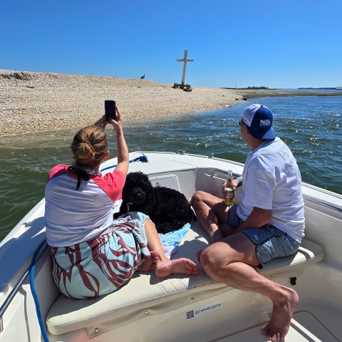Two people and a dog on a boat near a beach with a large cross.