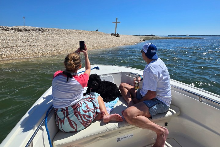Two people and a dog on a boat near a beach with a large cross.