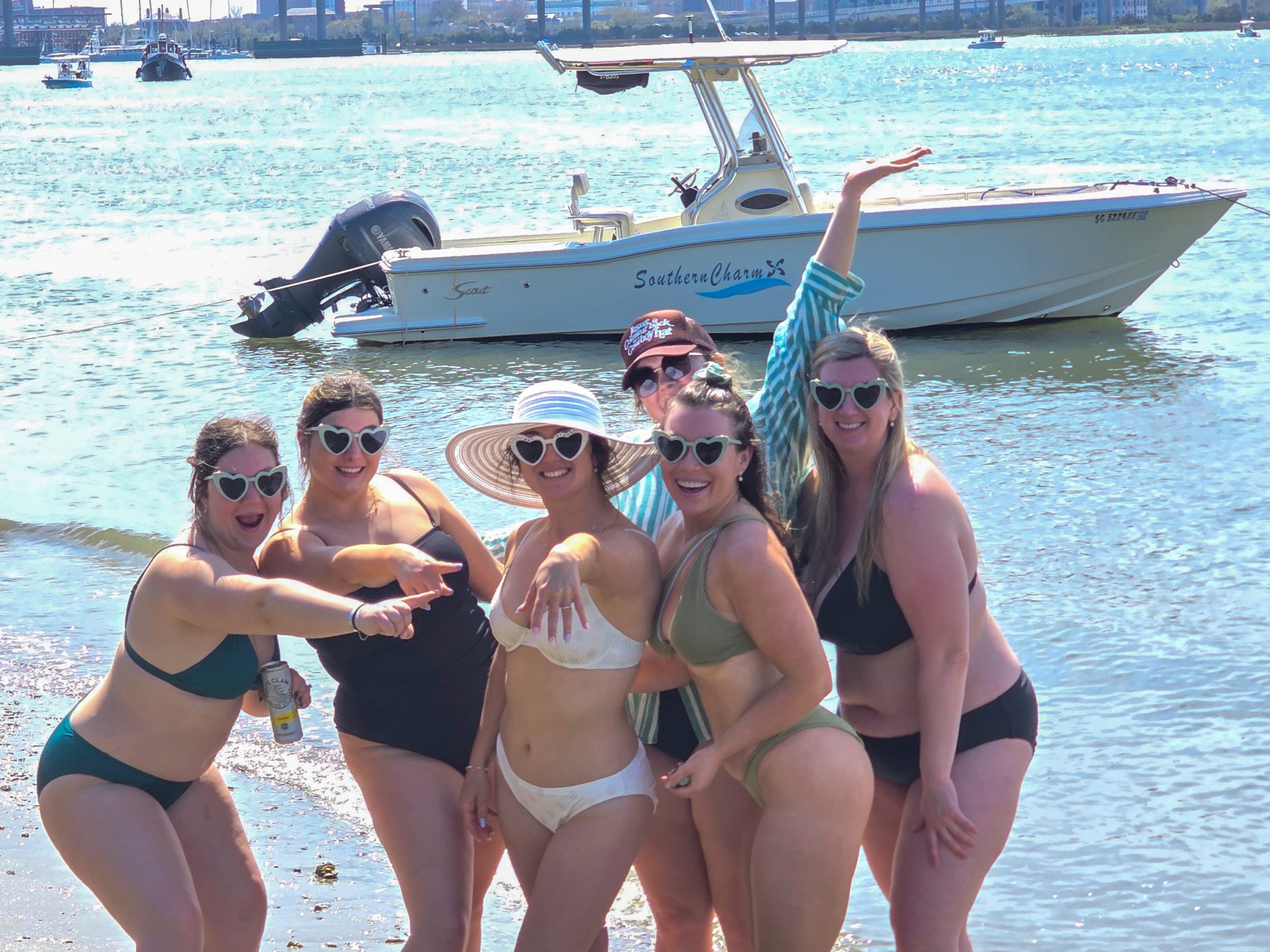 Group of six women in swimsuits posing by a boat on a sunny beach.