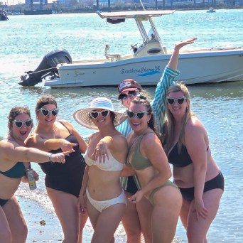 Group of six women in swimsuits posing by a boat on a sunny beach.