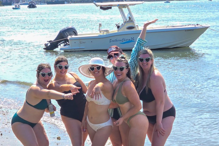 Group of six women in swimsuits posing by a boat on a sunny beach.