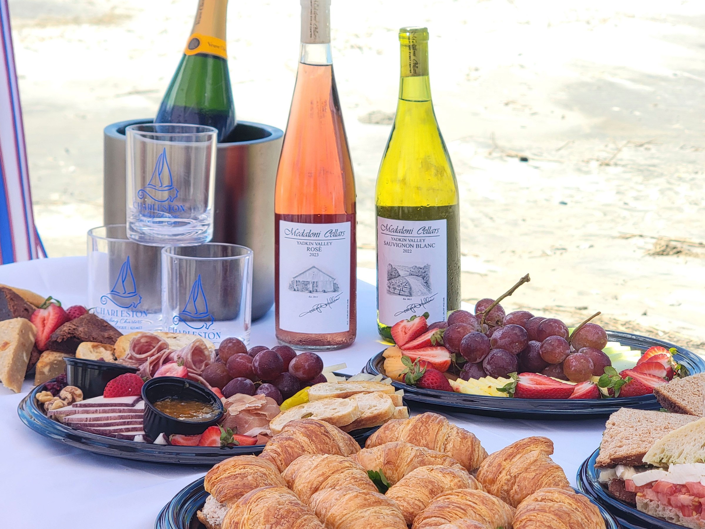 Picnic setup with croissants, sandwiches, wine bottles, and fruit platters on a beach background.