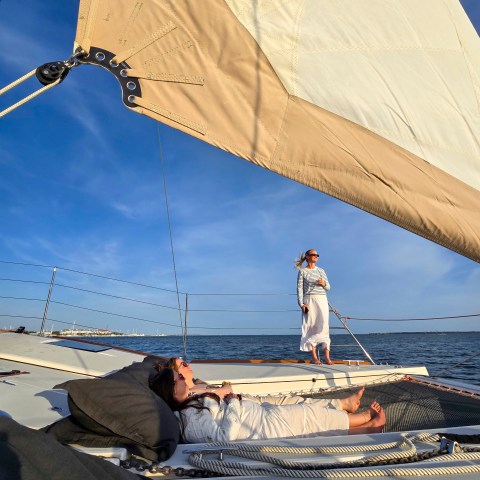 Two people relax on a sailboat, one lying down, another standing near the sail.