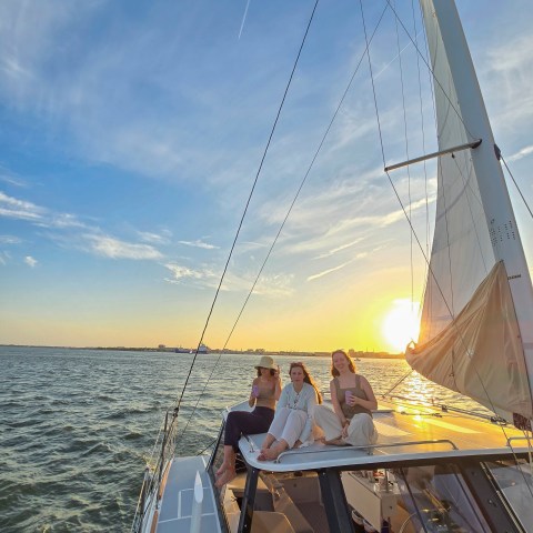Three people sitting on a sailboat at sunset, with a wide view of the ocean and sky.