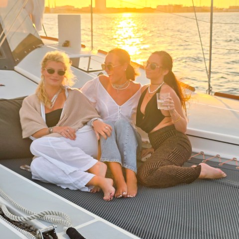 Three women sitting on a yacht deck at sunset, smiling and enjoying drinks.