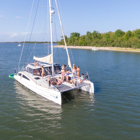 Group of people on a catamaran near a forested shoreline on a sunny day.