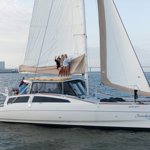 Catamaran with people on deck sailing near city waterfront and bridge.