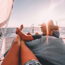 Two people relaxing on a sailboat with a sunset over the ocean.