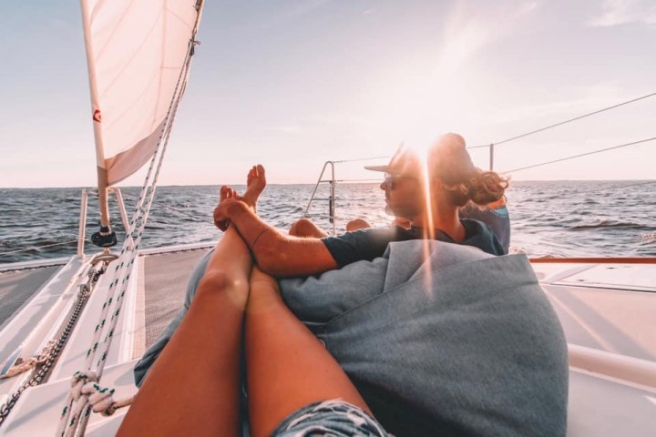 Two people relaxing on a sailboat with a sunset over the ocean.