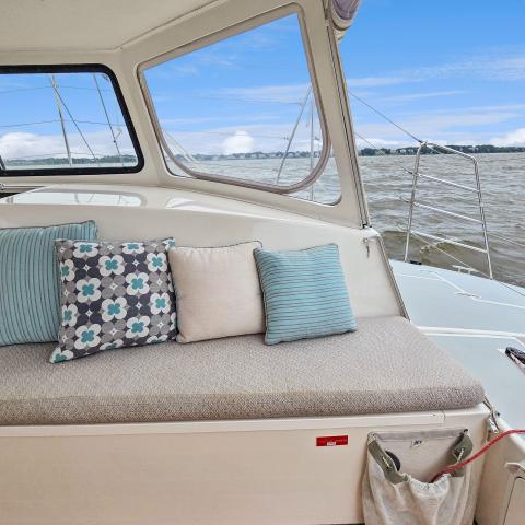 Boat interior with cushioned seating, decorative pillows, and view of water through windows.