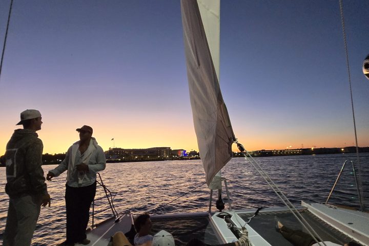 People on a sailboat at sunset with city lights in the distance.