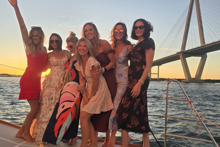 Group of women posing on a boat at sunset with a bridge in the background.